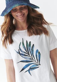 T-shirt blanche avec un imprimé de feuilles bleu et vert. Modèle portant un chapeau seau tissé bleu marine et vert. Tissu texturé visible.