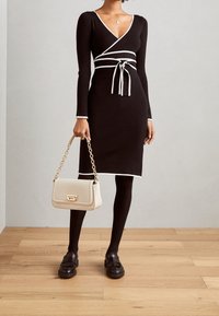 Robe noire côtelée avec bordure blanche, col en V, manches longues et taille nouée. Associée à un sac à bandoulière beige et des mocassins noirs.
