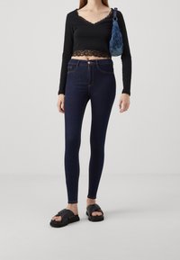 Mujer llevando vaqueros ajustados de color azul oscuro, top corto negro con encaje, sandalias negras de tira y cargando un bolso de hombro con un patrón azul.