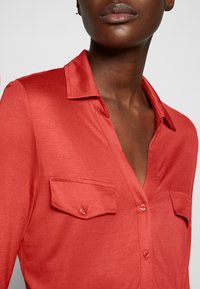 Camicia rossa con una texture liscia, colletto e chiusura con bottoni. Presenta due tasche sul petto e un design a scollo a V.