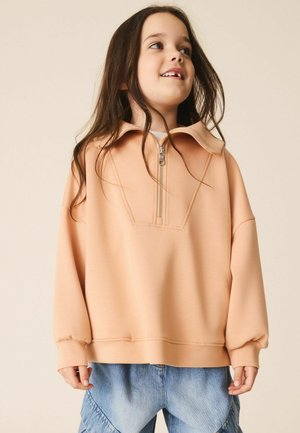 Young girl smiling wearing a peach half-zip sweatshirt and blue denim jeans against a plain beige background.