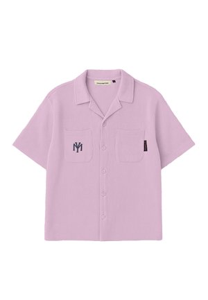 Jeune homme debout portant une chemise à manches courtes rose poussiéreux à boutons et un short assorti, des chaussettes blanches, des chaussures beiges sans lacets et une casquette bleu marine.