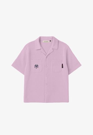 Junger Mann steht, trägt ein staubig rosa kurzärmeliges Hemd mit Knöpfen und passende Shorts, weiße Socken, beige Slipper und eine marineblaue Kappe.