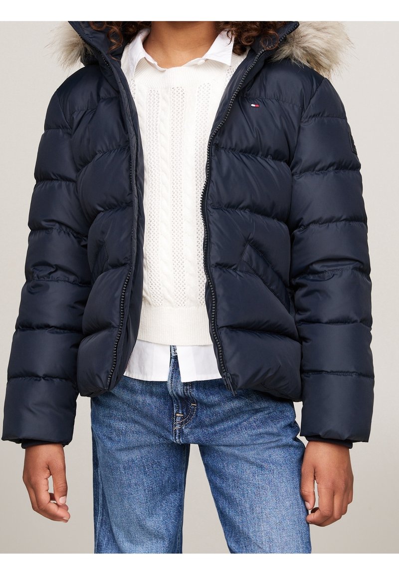 Enfant portant une doudoune bleu marine avec capuche bordée de fourrure, pull en maille blanche, chemise blanche et jean bleu sur un fond uni.