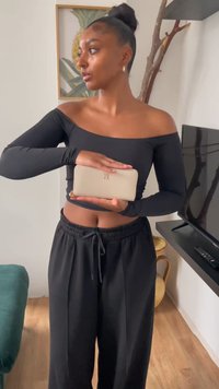 Krém színű pénztárca cipzárral, aranyszínű logóval, egy fekete hosszú ujjú felsőben és fekete nadrágban tartva, semleges háttér előtt.