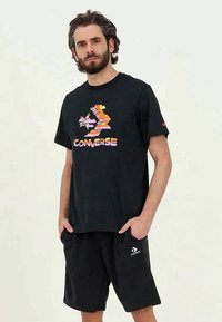 Vīrietis ar bārdu un cirtainiem matiem, ģērbies melnā Converse t-kreklā ar krāsainu logo un melnās šortās ar baltu Converse logo, rokas kabatās.