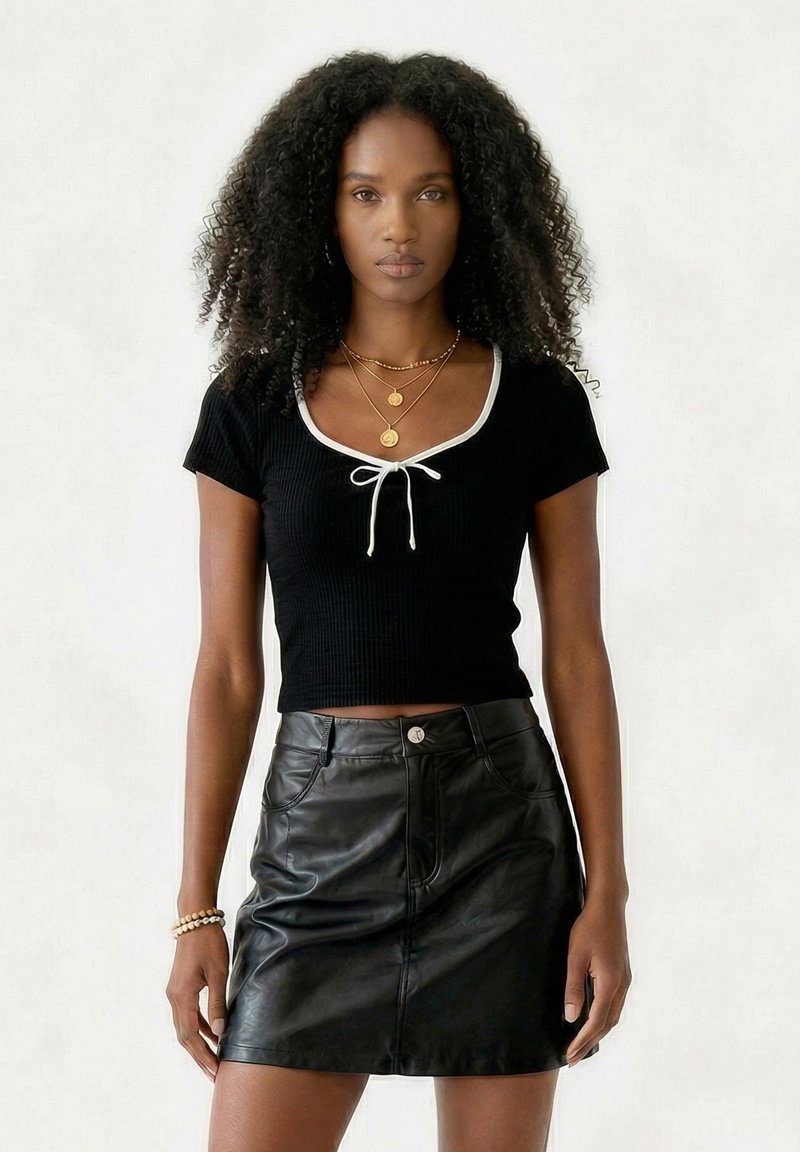 Femme aux cheveux bouclés portant un haut noir à manches courtes avec bordure blanche et une jupe en cuir noire, posant sur un fond clair uni.