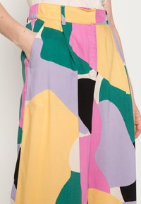Pantalon à jambes larges coloré présentant un mélange de formes abstraites vertes, violettes, roses, jaunes et noires. Tissu léger avec poches latérales.