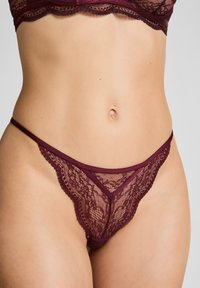 Vrouw in bordeauxrode kant beha en bijpassende kanten string, die haar middel en heupen toont tegen een effen achtergrond.