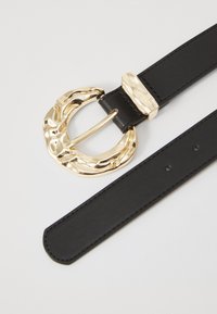Cinto de couro preto com uma grande fivela dourada, martelada, com um design curvado e um acento retangular em metal dourado.