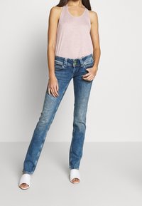 Haut sans manches rose pâle, jeans droit en denim bleu avec un délavage usé, et talons ouverts blancs. Bouton accent vert notable sur les jeans.