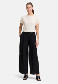 Lichtbeige top met korte mouwen, gecombineerd met hoge zwarte wijde broek. Model draagt zwarte hakken. Gladde stoftextuur.