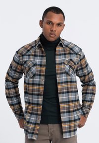 Geruit flanellen shirt in grijs, bruin en geel, met twee borstzakken en een knoopsluiting aan de voorkant. Gemaakt boven een donker coltrui.
