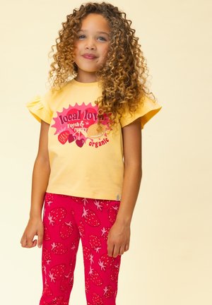 Meisje met krullend haar, gekleed in een geel shirt met ruches aan de mouwen en een rode fruitprint, en een rode broek met aardbeienpatroon, staande tegen een crèmekleurige achtergrond.