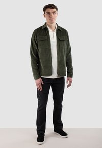 Grøn kordfløjljakke med to brystlommer, lynlås foran, båret over en hvid polo, parret med mørke jeans og sorte sneakers.