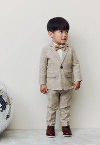 Costume à carreaux beige avec une veste et un pantalon assorti, chemise blanche et nœud papillon marron. Chaussures marron complètent la tenue. Boule disco argentée à proximité.