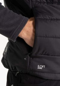 Black insulated vest with horizontal stitching, featuring a zippered pocket and "Iloft Ecofill" label. Soft fleece sleeve detail visible.