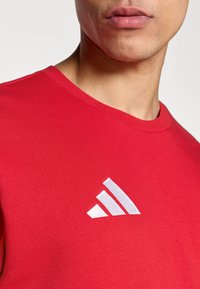 Κόκκινο βαμβακερό μπλουζάκι με στρογγυλή λαιμόκοψη και λευκό λογότυπο Adidas με τρεις ρίγες στην αριστερή πλευρά. Λεία  υφή.