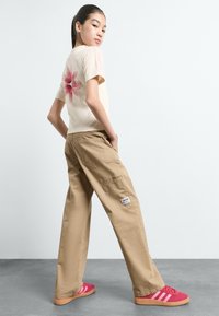 T-shirt bege com estampado floral rosa e três riscas rosas nas mangas, combinado com calças cargo bege largas e ténis vermelhos com riscas brancas.