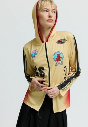 Lahka rumena jakna s kapuco in črnimi rokavi, zapenjanjem z zadrgo ter različnimi pisano obarvanimi našitki in grafičnimi motivi spredaj ter na rokavih.