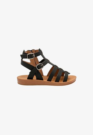 Sandale gladiateur en cuir noir avec semelle plate beige et deux boucles argentées réglables sur les brides de cheville.