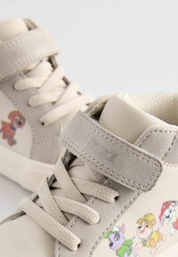 Beżowe buty typu high-top wykonane z materiałów tekstylnych i syntetycznych. Posiadają sznurowany design, rzep oraz kolorowe nadruki postaci z kreskówek.