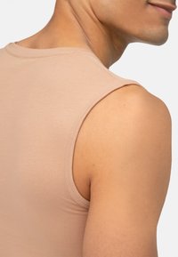 Maglietta senza maniche di colore nude realizzata in tessuto morbido, con scollatura arrotondata e cuciture pulite lungo le aperture delle maniche e l'orlo. Colore simile alla pelle.