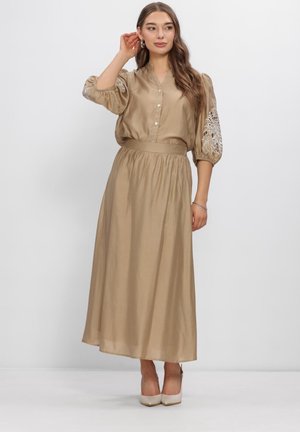 Vrouw draagt een beige satijnen blouse met bloemenborduursel op de mouwen en een bijpassende lange rok, poseert met hand bij het oor en lichte hakken.