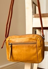 Gul läder crossbody-väska med framficka med dragkedja hängande på en vit trästol i ett neutralt rum.