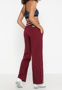 Bordowe spodnie dresowe z paskiem w paski i bocznymi guzikami, wykonane z gładkiego materiału. W połączeniu z czarnym sportowym biustonoszem i białymi butami.
