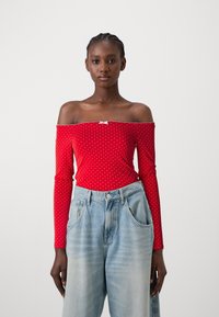Crvena off-shoulder majica dugih rukava s bijelim točkicama. Ima blagi rastezljiv materijal i mali dekorativni detalj na izrezu. Uparena s svijetloplavim širokim trapericama.