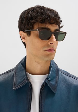 Giovane uomo che indossa occhiali da sole scuri, una camicia bianca e una giacca di pelle blu, che guarda leggermente di lato su uno sfondo semplice.