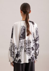 Donna con capelli lunghi legati che indossa una blusa bianca ampia con stampa di un paesaggio urbano architettonico nero, vista da dietro su uno sfondo semplice.