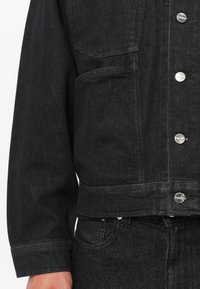 Chaqueta de mezclilla negra con botones a presión plateados, dos bolsillos frontales y un diseño ligeramente cortado. La tela tiene una textura y peso sutiles.