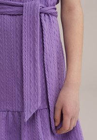 Levandų suknelė su tekstūruotu bangų raštu ir surištos juostos akcentu. Suknelė turi sluoksniuotą dizainą su platėjančiu kraštu ir minkšta, lengva medžiaga.