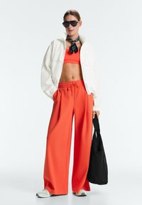 Femme portant une veste blanche oversize sur un crop top orange et un pantalon large orange, écharpe noire, lunettes de soleil, baskets blanches, tenant un sac cabas noir.