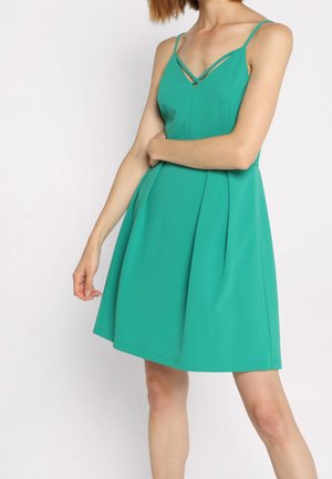 Femme portant une robe sans manches turquoise avec de fines bretelles et une jupe évasée, posant avec un bras croisé et l'autre pendant le long du corps.