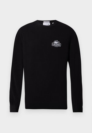 Двама души, носещи черни суичъри Lacoste; единият със светлосини дънки и шапка, другият с бежови къси панталони и бели маратонки, стоящи един до друг.