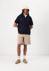 Camisa de punto marina de manga corta con cierre de botones, combinada con pantalones cortos beige y zapatos color beige claro. Calcetines blancos visibles. Atuendo casual de verano.