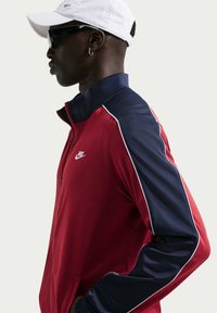 Κόκκινο και ναυτικό μπλε αθλητικό μπουφάν Nike με ψηλό γιακά, κλείσιμο με φερμουάρ και λευκές λεπτομέρειες. Διαθέτει λογότυπο στο στήθος και υφασμένα μανίκια.