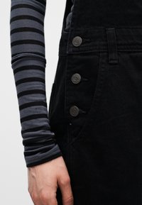 Κοντινή λήψη ενός χεριού δίπλα σε μαύρη φόρμα με τρία μεταλλικά κουμπιά και μια γκρι και μαύρη ριγέ μακρυμάνικη μπλούζα.