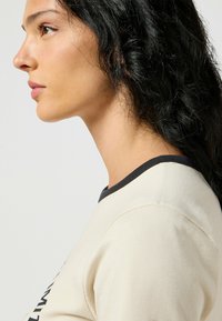 Top beige con escote oscuro, mangas cortas y tela ligera. La cabeza del modelo está girada, mostrando un perfil suave y cabello negro texturizado.