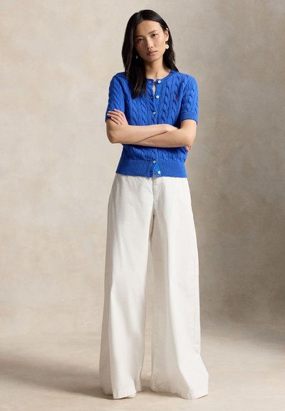 Mlada ženska z dolgimi temnimi lasmi stoji z rokami prekrižanimi, oblečena v modri kratek rokav pleteni pulover in široke bele hlače.