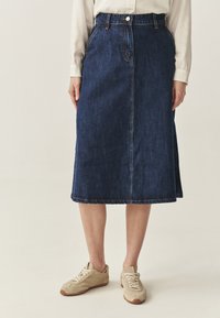 Falda midi de mezclilla en azul oscuro con corte recto, pasantes para cinturón, cierre delantero con botones y detalles de costura visibles. Combinada con zapatillas beige.