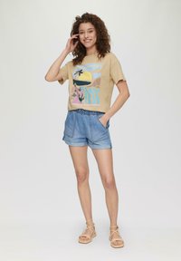 Beige T-Shirt mit buntem Grafikdesign, das einen Sonnenuntergang und Palmen zeigt, kombiniert mit hellblauen Denim-Shorts mit elastischem Bund.
