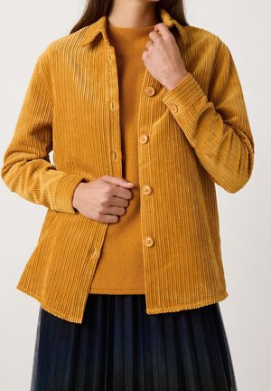 Persona con camisa de pana amarilla mostaza abotonada sobre suéter de punto a juego con falda plisada oscura, ajustando el cuello con la mano derecha.