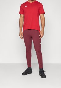 Rode sportieve t-shirt met korte mouwen en V-hals; gecombineerd met bordeauxkleurige slim-fit leggings en zwarte sportschoenen. Heeft het Adidas-logo.