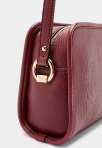 Wijnkleurige leren handtas met een gladde textuur, goudkleurige hardware en een ritssluiting. Heeft een ronde vorm en een zijriem.