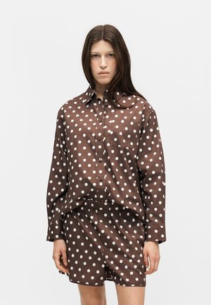 Jeune femme portant une chemise marron à manches longues avec col et un short assorti à pois blancs, debout devant un fond uni.