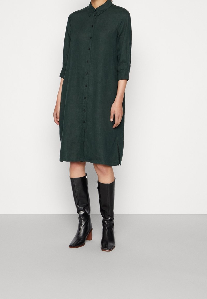 Femme portant une robe-chemise vert foncé à manches longues avec des bottes en cuir noires montant jusqu'aux genoux sur un sol gris.